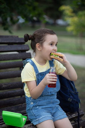 Cute little schoolgirl eating from lunch box outdoor sitting on a bench. Food for kids. School conceptの写真素材