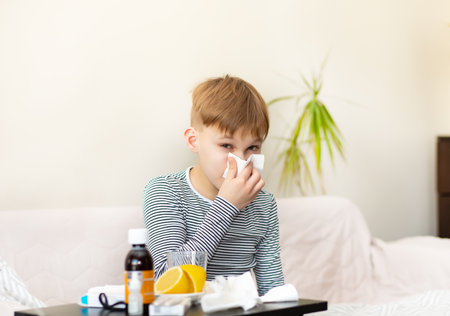 Sick little kid boy sits on bed at home and blows his nose. Flu season. Healthcare conceptの写真素材
