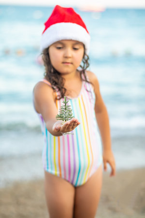 Cute little girl standing on the beach in Santa hat and striped swimsuit and smiling. Kid holding Christmas tree toy in miniature. Christmas tropical vacation conceptの写真素材