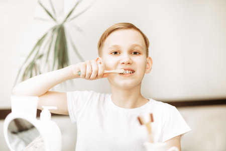Smiling blonde teenager kid boy brushing teeth at home looking in small mirror. Morning routineの写真素材