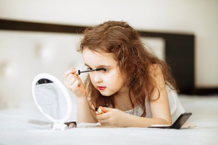 Kids makeup. Happy cute girl doing makeup lies on bed in the bedroom. Kid using mascara looks in the mirror. Skin care concept. Cosmetics for childrenの写真素材