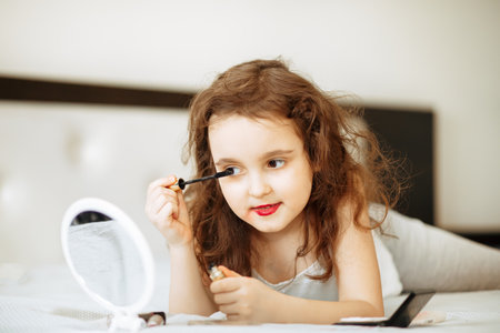 Kids makeup. Happy cute girl doing makeup lies on bed in the bedroom. Kid using mascara looks in the mirror. Skin care concept. Cosmetics for childrenの写真素材