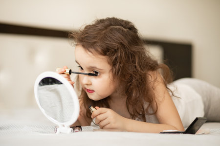 Kids makeup. Happy cute girl doing makeup lies on bed in the bedroom. Kid using mascara looks in the mirror. Skin care concept. Cosmetics for childrenの写真素材