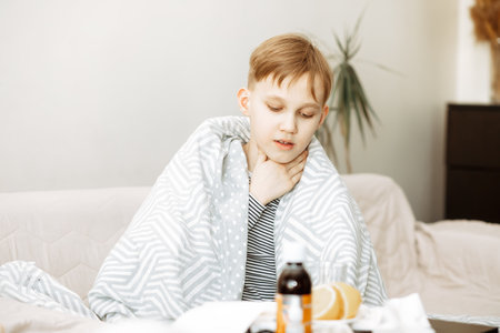 Sick little teen boy sits on bed at home and holding his sore throat. Flu season. Healthcare conceptの写真素材
