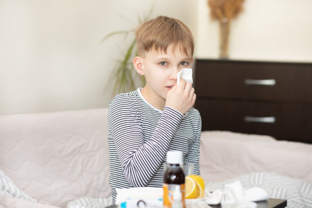 Sick little kid teen boy sits on bed at home and blows his nose. Flu season. Healthcare conceptの写真素材