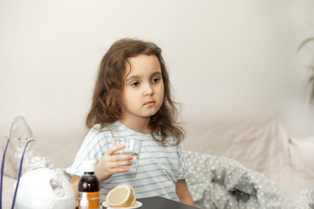Sick sad little kid girl sits on bed at home, holding glass of water and is going to take medication. Flu season. Healthcare conceptの写真素材