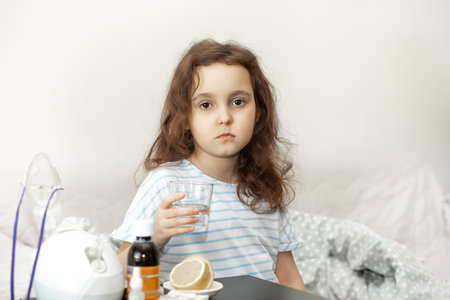 Sick sad little kid girl sits on bed at home, holding glass of water and is going to take medication. Flu season. Healthcare conceptの写真素材