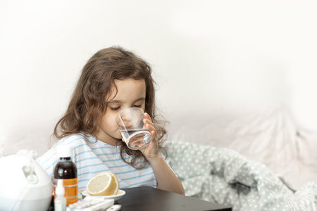 Sick sad little kid girl sits on bed at home, drinking water with medication. Flu season. Healthcare conceptの写真素材