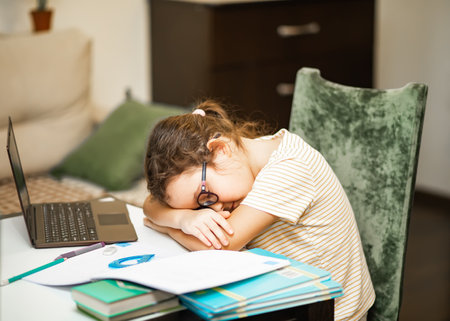 Little cute girl pupil at home making homework. Kid sleeping resting head on books. Child studying online on laptop. Kid educationの写真素材