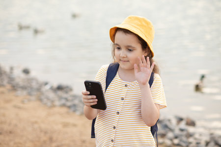 Cute little girl in yellow hat taking selfie with mobile phone outdoor in a park near by lake. Kid blogger recording video vlog.の写真素材