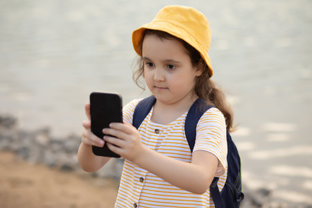 Cute little girl in yellow hat taking selfie with mobile phone outdoor in a park near by lake. Kid blogger recording video vlog.の写真素材
