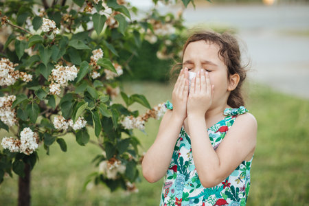Little kid girl with seasonal allergy gets cold and blows her nose outdoor nearby blooming trees. Healthcare, pollen and flowering allergy in spring concept. World Allergy Dayの写真素材