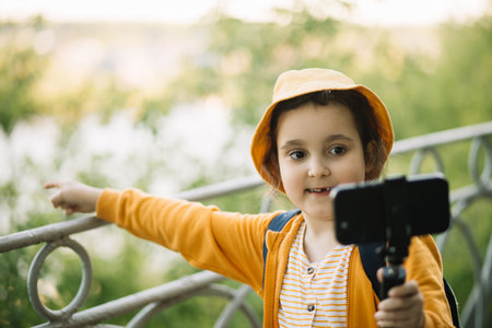 Cute little girl in yellow hat taking selfie with mobile phone outdoor in a park. Kid blogger recording video vlog.の写真素材