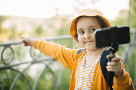 Cute little girl in yellow hat taking selfie with mobile phone outdoor in a park. Kid blogger recording video vlog.の写真素材