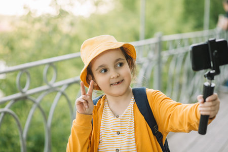 Cute little girl in yellow hat taking selfie with mobile phone outdoor in a park. Kid blogger recording video vlog.の写真素材