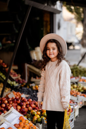 Cute little girl in a hat with net bag buying fruits and vegetables at organic farmers market. Green and healthy eating lifestyle. Enjoying shopping for healthy food.の写真素材