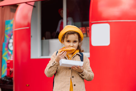 Cute little girl eating hot dog nearby food truck on a street. Food delivery or take away food conceptの写真素材