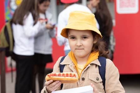 Cute little girl eating hot dog nearby food track on a street. Food delivery or take away food conceptの写真素材