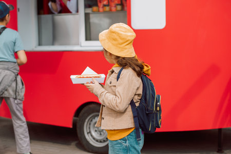 Cute little girl eating hot dog nearby food truck on a street. Food delivery or take away food conceptの写真素材
