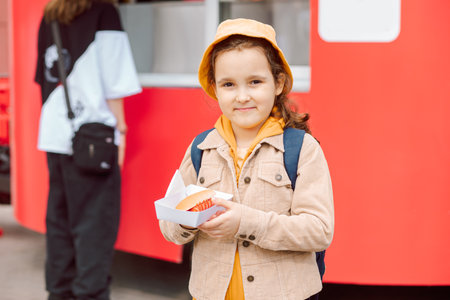 Cute little girl eating hot dog nearby food track on a street. Food delivery or take away food conceptの写真素材