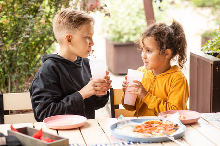Cute girl and teenager boy brother and sister eat pizza lunch and drinking lemonade in outdoor cafe and having fun. Happy family.の写真素材