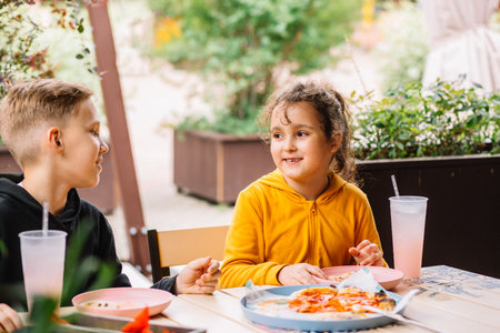 Cute girl and teenager boy brother and sister eat pizza lunch and drinking lemonade in outdoor cafe and having fun. Happy family.の写真素材