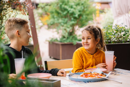 Cute girl and teenager boy brother and sister eat pizza lunch and drinking lemonade in outdoor cafe and having fun. Happy family.の写真素材