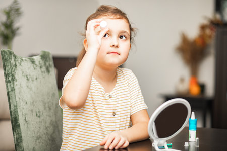 Cute little girl uses moisturizing drops inserting contact lenses into her eye at home. Vision correction and ophthalmology for childrenの写真素材