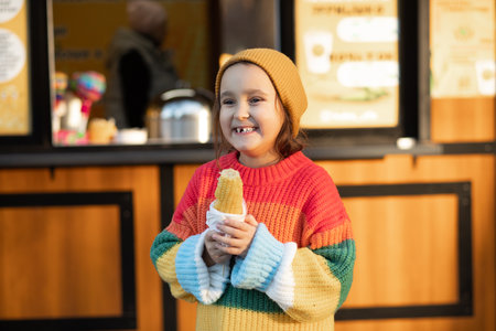Little cute girl eating delicious tasty corn on the cob in city parkの写真素材