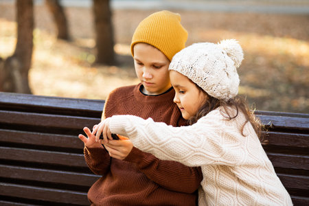 Kids girl and boy play online games or read social networks on mobile smart phones. Children sitting on bench outdoor in parkの写真素材