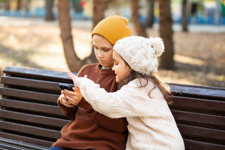 Kids girl and boy play online games or read social networks on mobile smart phones. Children sitting on bench outdoor in parkの写真素材