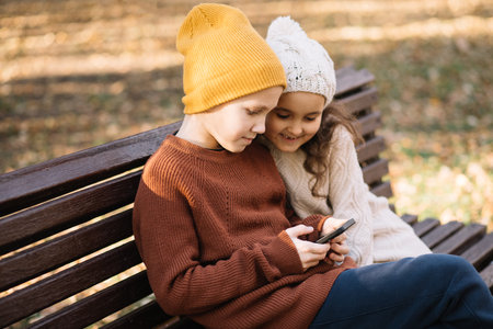 Kids girl and boy play online games or read social networks on mobile smart phones. Children sitting on bench outdoor in parkの写真素材