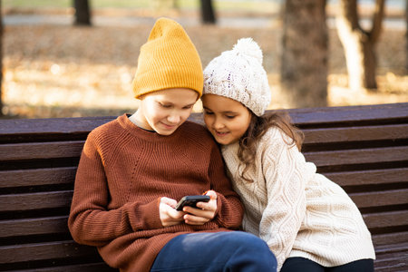 Kids girl and boy play online games or read social networks on mobile smart phones. Children sitting on bench outdoor in parkの写真素材