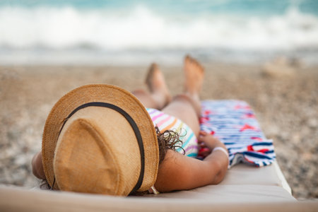 Cute little girl in a hat and striped swimsuit lies on a sun lounger and sunbathes on the beach in summer. Holiday vacation with childrenの写真素材