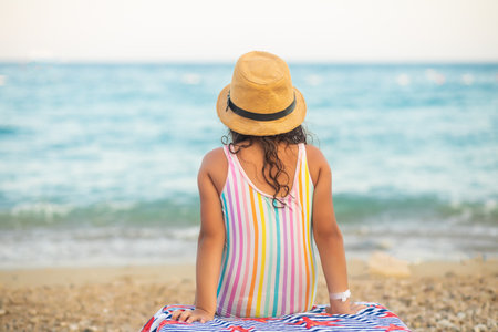 Cute little girl in a hat and striped swimsuit sitting on the beach in summer. Holiday vacation with childrenの写真素材