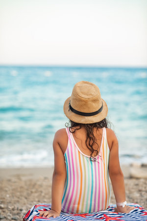 Cute little girl in a hat and striped swimsuit sitting on chaise lounge on the beach in summer. Holiday vacation with childrenの写真素材