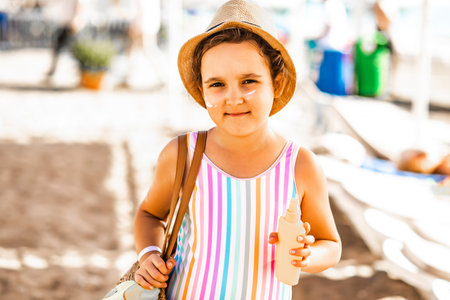 Cute little girl in a hat, striped swimsuit holding bottle of sunscreen in a hand at the beach in summer. Holiday vacation with childrenの写真素材