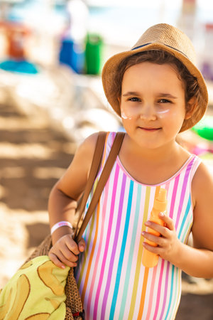 Cute little girl in a hat, striped swimsuit holding bottle of sunscreen in a hand at the beach in summer. Holiday vacation with childrenの写真素材