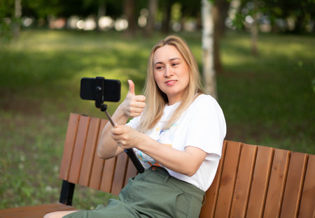 Pretty woman taking selfie with mobile phone looking at camera in a park. Blogger recording video vlogの写真素材