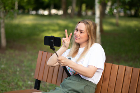 Pretty woman taking selfie with mobile phone looking at camera in a park. Blogger recording video vlogの写真素材