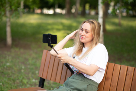 Pretty woman taking selfie with mobile phone looking at camera in a park. Blogger recording video vlogの写真素材