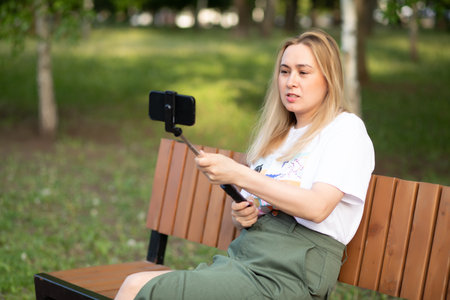 Pretty woman taking selfie with mobile phone looking at camera in a park. Blogger recording video vlogの写真素材