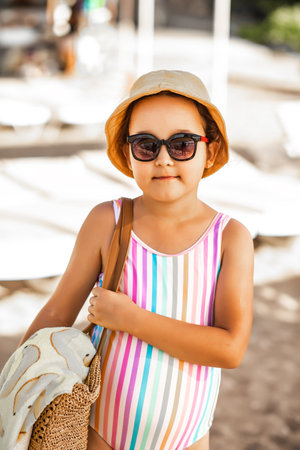 Cute little girl in striped swimsuit, hat and sunglasses standing at the beach in summer. Holiday vacation with children.の写真素材