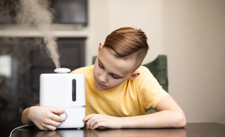 Cute teenager kid boy sitting near a humidifier with outgoing steam in a roomの写真素材