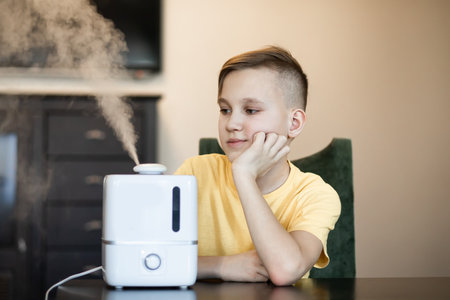 Cute teenager kid boy sitting near a humidifier with outgoing steam in a roomの写真素材