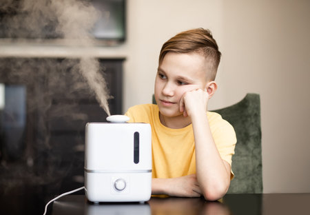 Cute teenager kid boy sitting near a humidifier with outgoing steam in a roomの写真素材