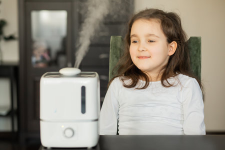 Cute little kid girl sitting near a humidifier with outgoing steam in a room.の写真素材