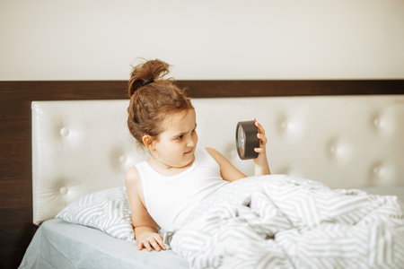 Cute little kid girl sitting on bed waking up in the morning and holding alarm clock and smilingの写真素材