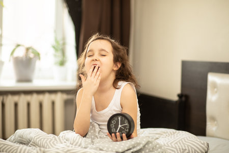 Cute little kid girl sitting on bed waking up in the morning and holding alarm clock and smilingの写真素材