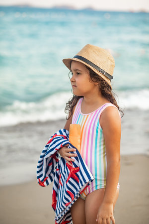 Cute little girl in a hat, striped swimsuit holding bottle of sunscreen and towel in a hand at the beach in summer. Holiday vacation with childrenの写真素材